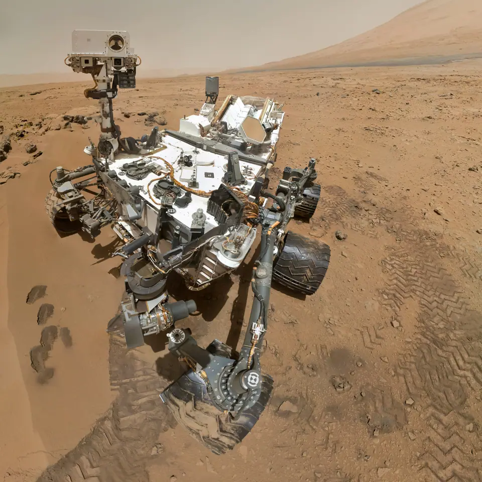 Autorretrato del vehículo Curiosity sobre el suelo rojizo de Marte, con huellas de sus ruedas y el monte Sharp al fondo bajo un cielo pálido