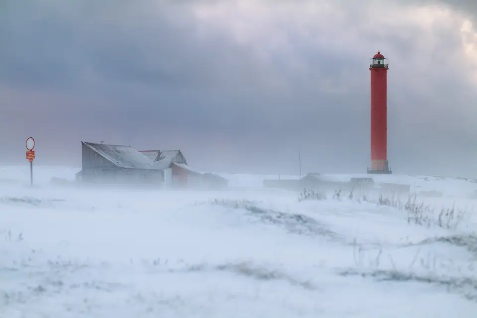 Faro rojo y casas bajo una ventisca, con la nieve arrastrada por el viento