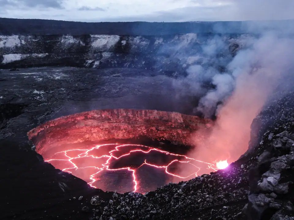 Lago de lava en el cráter Halemaʻumaʻu del Kīlauea: superficie roja incandescente cruzada por grietas brillantes y una columna de gases que sube desde el borde