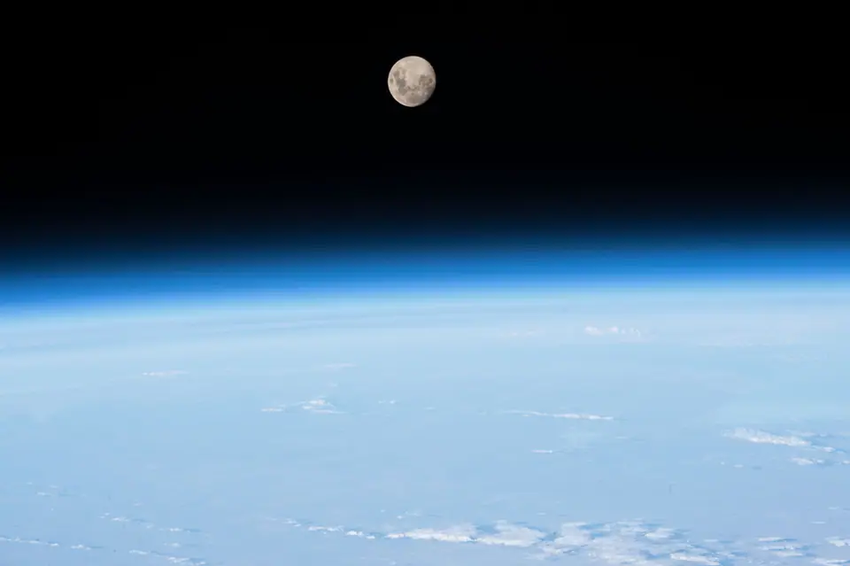 El borde de la Tierra visto desde la Estación Espacial Internacional: la atmósfera forma una franja azul delgada entre las nubes y el negro del espacio, con la Luna arriba