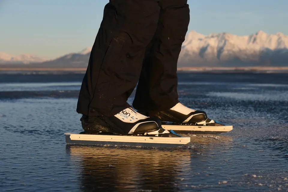 Patines de hoja larga apoyados sobre el hielo liso de un lago congelado, con montañas nevadas al fondo