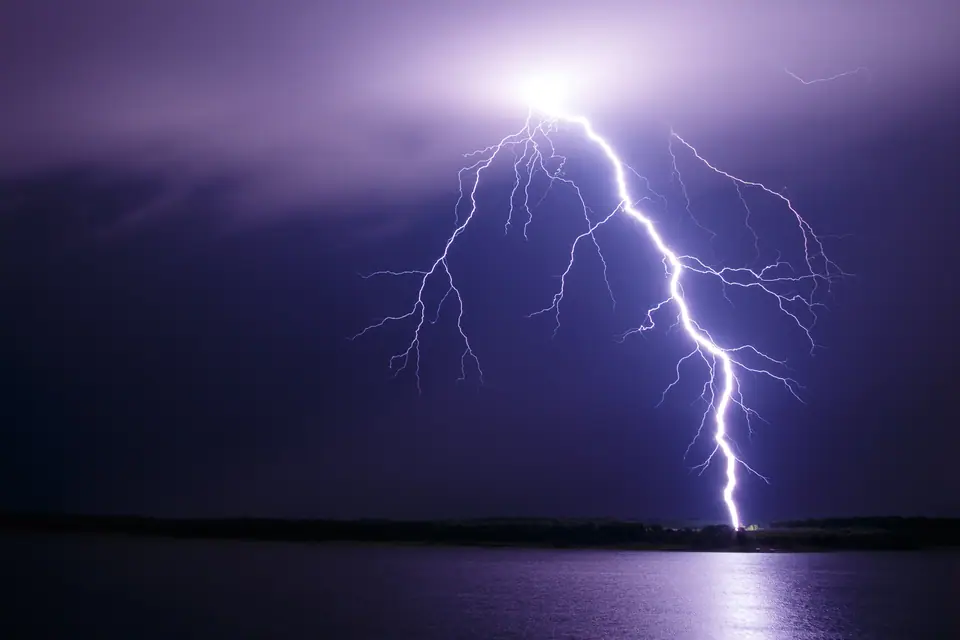 Un rayo ramificado baja de la nube y toca tierra en la orilla de un lago, de noche, con varias ramas laterales que no llegan al suelo
